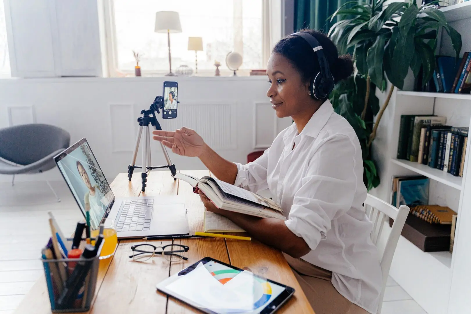 A woman is sitting at her desk with headphones on and a book.